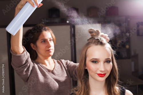 hairdresser fixing a coiffure with an unusual bun of a young beautiful girl using a hair spray in a beauty salon. concept of professional stylist studying © luckyphoto