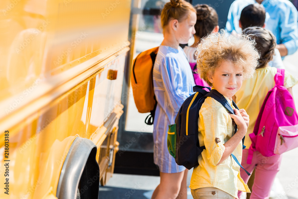 adorable curly schoolboy with his classmates standing near school bus