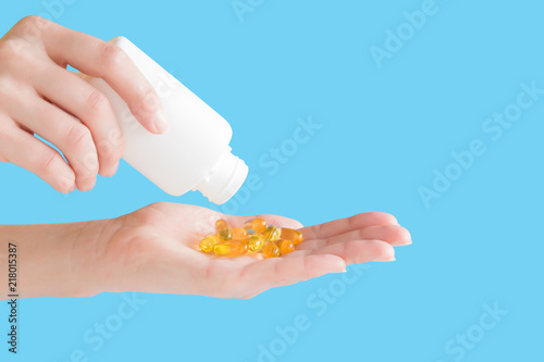 Woman spilling out fish oil capsules from bottle in her hand. Receiving vitamins. Nutritional supplements and healthcare concept. Copy space. Empty place for text or logo on blue background.