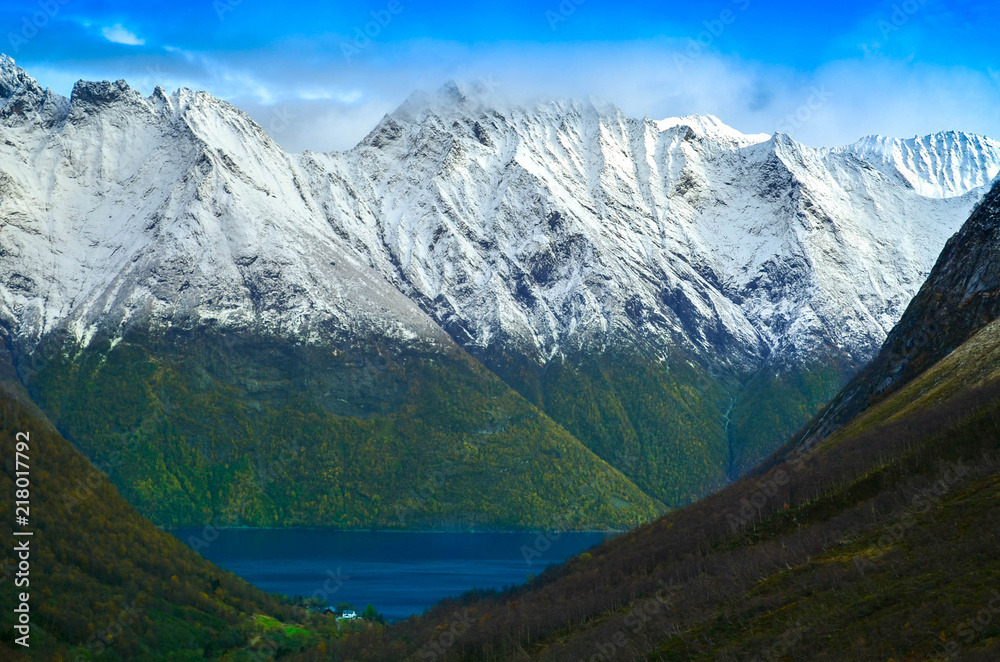 Group of hikers walking in the mountains. A small wooden cabin, tents and holiday makers in the alley in the middle of mountains, next to Fjords. End of autumn, cold weather, bonfire and snowy peaks