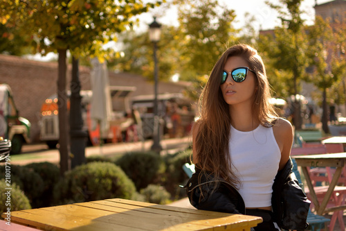 mujer joven atractiva  con gafas de sol y camiseta blanca sentada en una silla de un parque en una calle de la ciudad