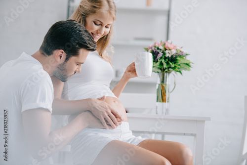 smiling pregnant woman with cup of tea resting on chair while husband touching belly near by at home