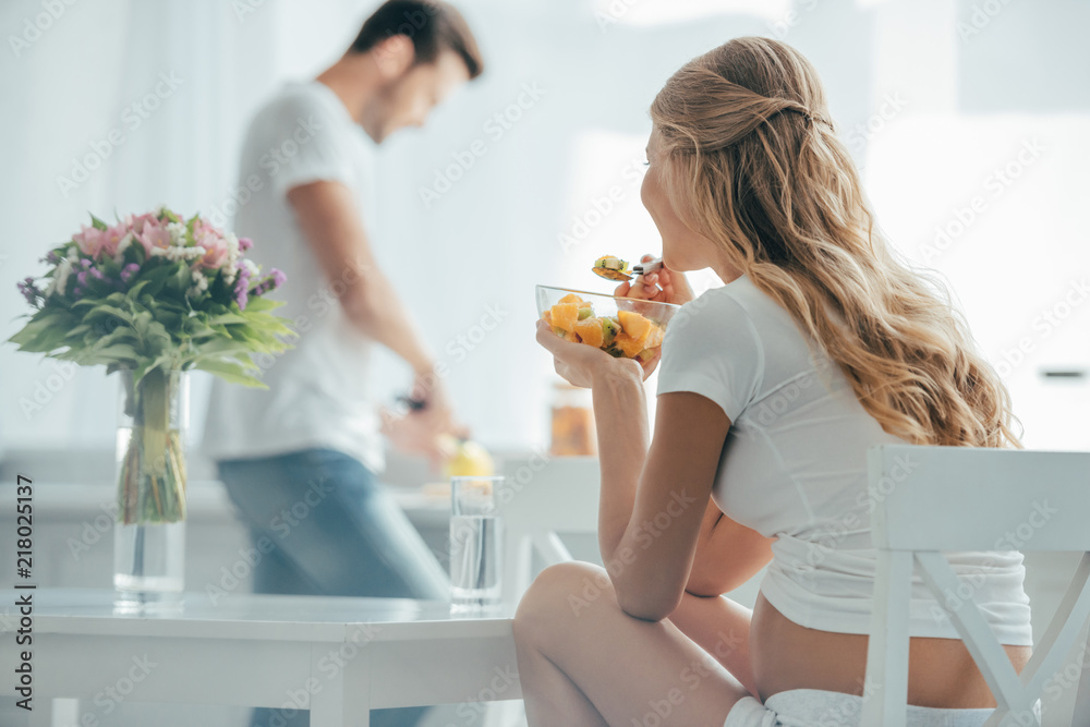 selective focus of pregnant woman eating fruits salad while husband standing at counter in kitchen selective focus of pregnant woman eating fruits salad while husband standing at counter in kitchen