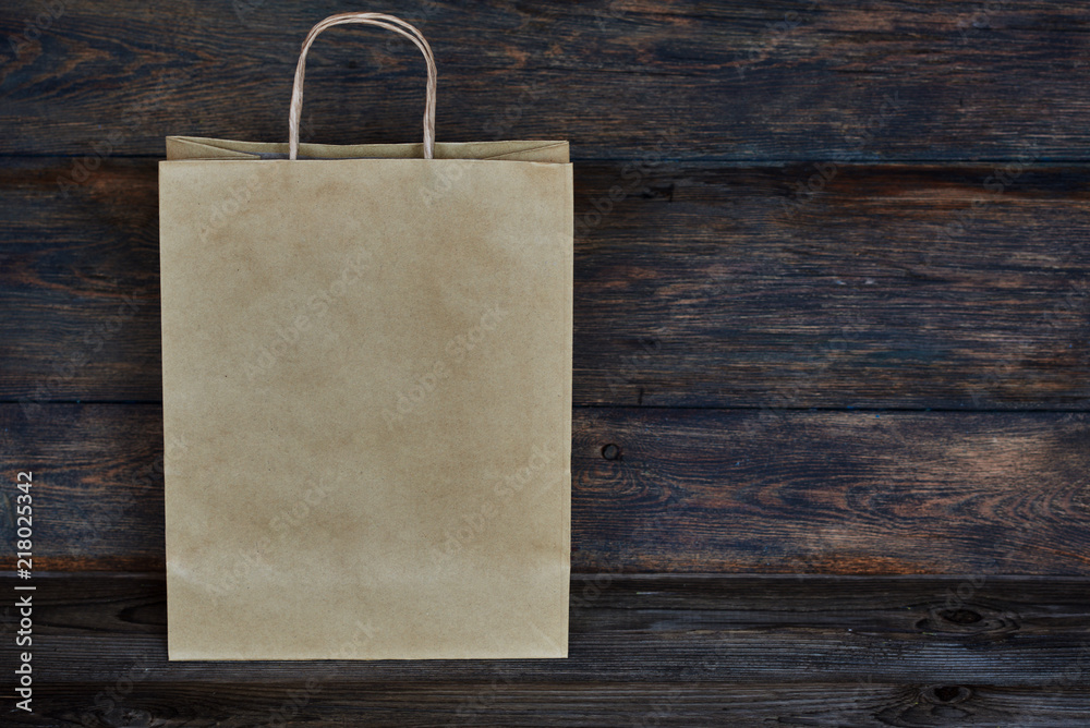 Mock-up of brown craft paper package with handles, empty shopping bag with area for your logo or design, wooden board in the background, food delivery, sale, consumerism and advertising concept. Mock-up of brown craft paper package with handles, empty shopping bag with area for your logo or design, wooden board in the background, food delivery, sale, consumerism and advertising concept.