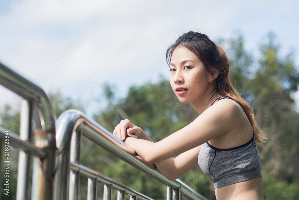 Young woman relaxing after jogging exercise on fence at park to freshen her body and enjoy warm light in morning. Young asian woman take a break from running execise. Outdoor exercise activity.