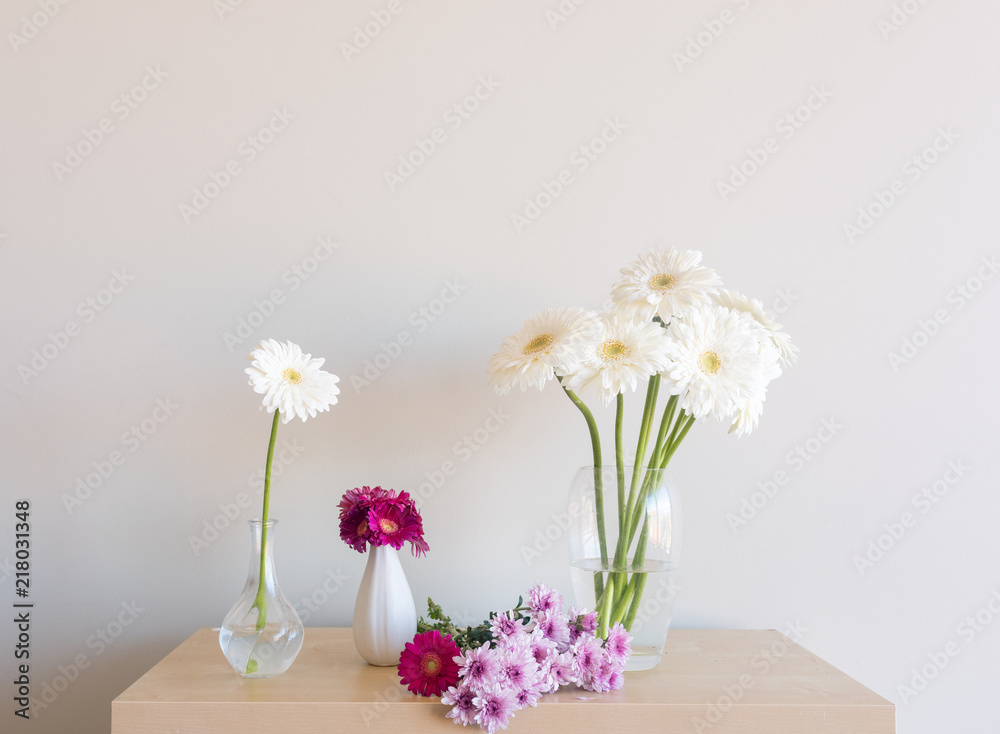 White and pink gerberas and purple chrysanthemums in vases and on wooden shelf against neutral wall background (selective focus
