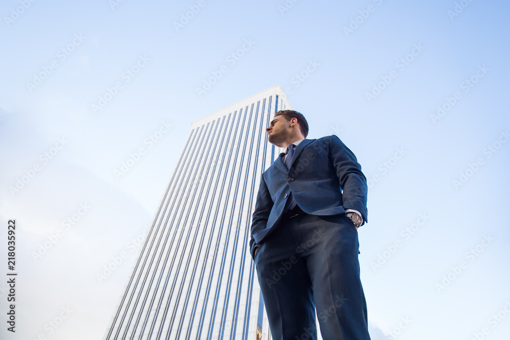 From below view of stylish man in elegant suit posing confidently on background of modern office skyscraper From below view of stylish man in elegant suit posing confidently on background of modern office skyscraper