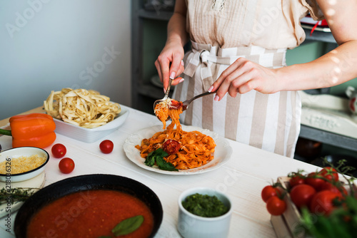 Chef in the kitchen holding a fork with pasta and sauce. Food ingredients and utensils on background. Pasta with pesto sauce, basil, cherry-tomatoes, garlic and thyme on old grunge white table.