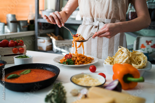 Woman in kitchen making delicious pasta with sauce using a pan at home. Italian rural cooking still life. Wooden board, fresh vegetables, cooked pasta and a pan of sauce on a wooden kitchen table.