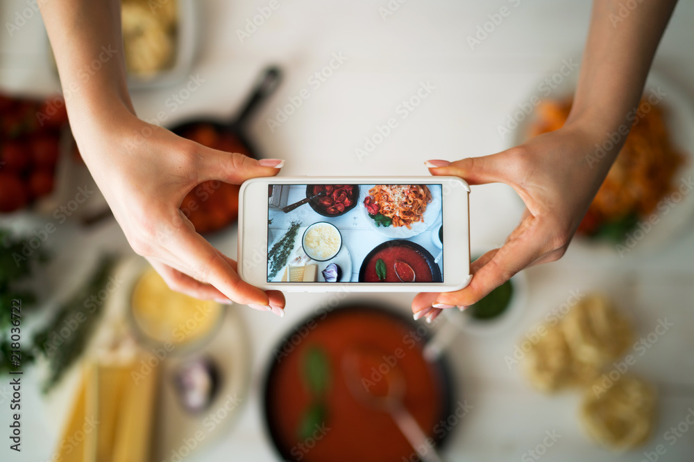 Hands with the smart phone pictures of meal. Young woman, cooking blogger is cooking at the home kitchen in sunny day and is making photo at smartphone. Instagram food blogger workshop concept. - obrazy, fototapety, plakaty 