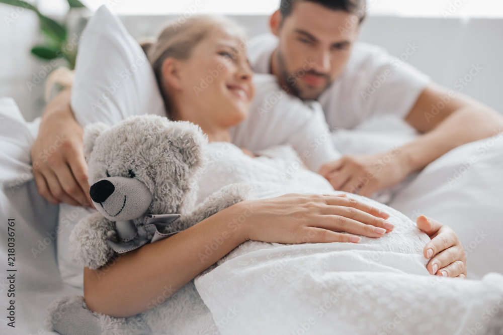 pregnant woman in white nightie with teddy bear on sofa and husband near by at home pregnant woman in white nightie with teddy bear on sofa and husband near by at home