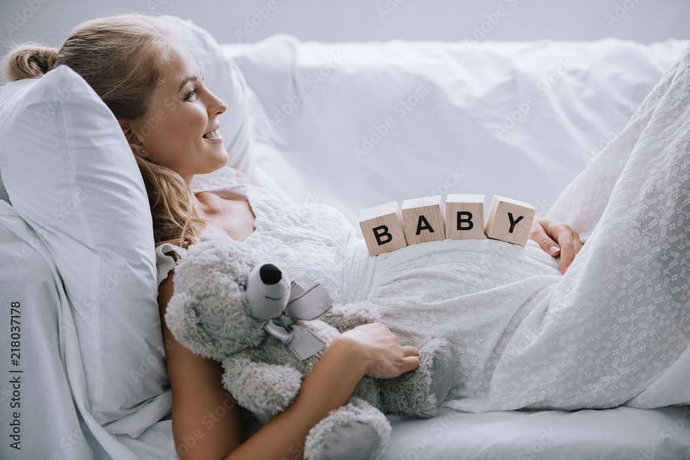 side view of smiling pregnant woman in white nightie with teddy bear and wooden blocks with baby lettering on belly resting on sofa side view of smiling pregnant woman in white nightie with teddy bear and wooden blocks with baby lettering on belly resting on sofa