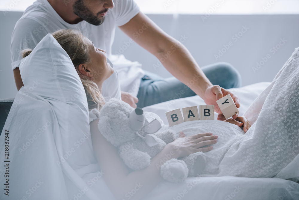 Obraz premium partial view of man putting wooden blocks with baby lettering on belly of pregnant wife with teddy bear at home