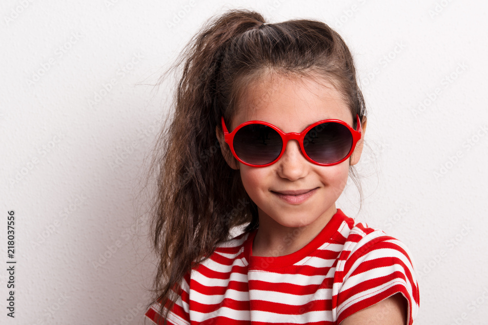 A small girl with red sunglasses and striped T-shirt standing in a studio.