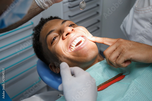 Happy young African-American man in chair of dentist pointing at hurtful tooth in mouth while doctor examining cavity