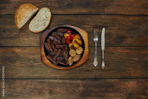 On a dark wooden table is a cast-iron pan with slices of juicy meat and grilled vegetables, fresh bread is nearby