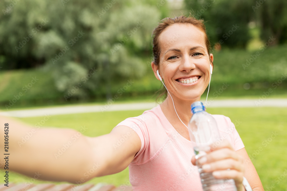 fitness, sport and healthy lifestyle concept - happy woman in earphones and water taking selfie in park fitness, sport and healthy lifestyle concept - happy woman in earphones and water taking selfie in park