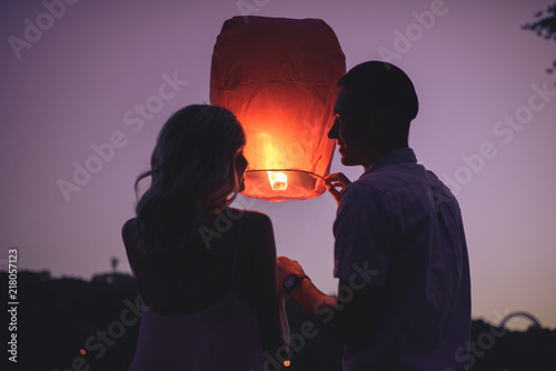 silhouettes of couple launching sky lantern on river beach in evening and looking at each other photo