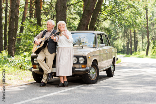 senior wife with husband playing guitar leaning on car on road in forest © LIGHTFIELD STUDIOS