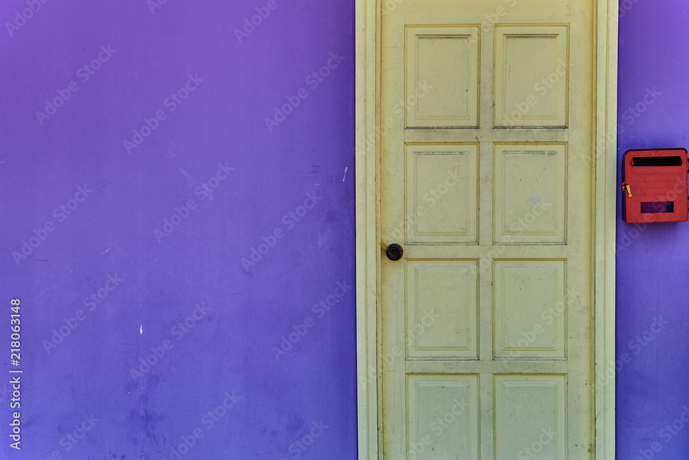 An old shabby light yellow door with a bright red mailbox against the background of a purple wall. Abstract background of the front of the building with a free place for text.