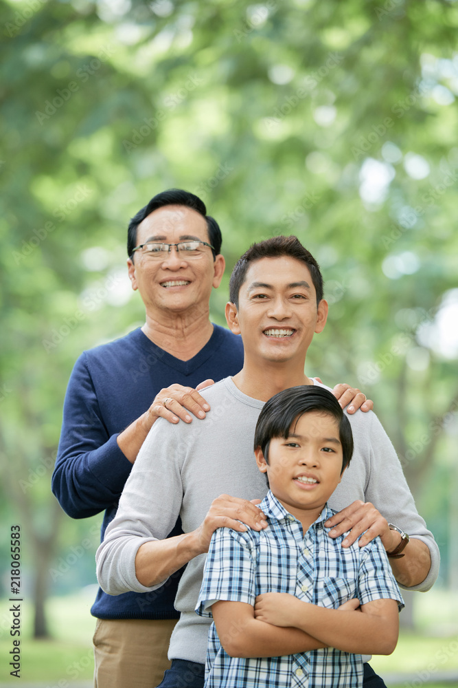 Cute Asian boy smiling and keeping arms crossed while standing near cheerful father and grandfather on blurred background of park Cute Asian boy smiling and keeping arms crossed while standing near cheerful father and grandfather on blurred background of park