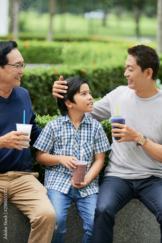 Adorable Asian boy holding cup of yummy drink and smiling while sitting at park near father and grandfather