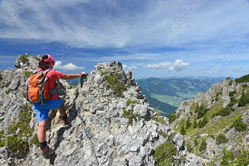 Female hiker climbing at an exposed ridge close to the village Oberstdorf in the Allgau Alps, Bavaria, Germany. Alpine landscape with rocky mountains, forests and blue sky.