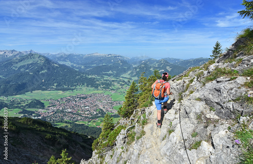 Rear view of a female hiker at an exposed spot at the Rubihorn mountain above the village Oberstdorf. Allgau Alps, Bavaria, Germany. Alpine landscape with rocky mountains, forests and blue sky.