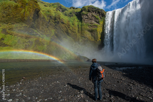 Urlauberin mit Rucksack, stehend vor Wasserfall auf Island, im Wassernebel hat sich ein Regenbogen gebildet