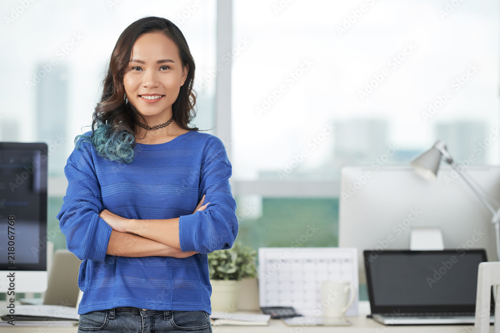 Lovely Asian female keeping arms folded and smiling while standing near desk in modern office