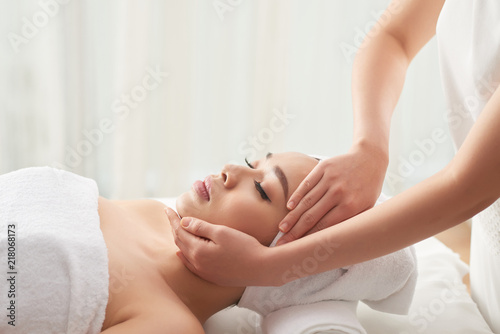 Crop hands of massage therapist rubbing face of pretty female client while working in spa salon