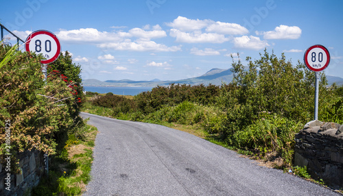 Country road with tall bushes along the sides, signs indicating an 80km speed limit, with mountains and ocean in the distance. Taken on a sunny summer day in Renvyle Ireland