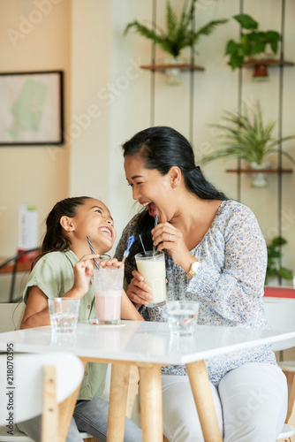 Adult Asian woman and adorable girl laughing and looking at each other while drinking delicious milkshakes in cozy cafe