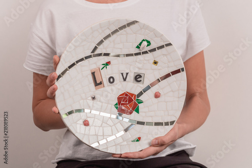 Young blonde woman in a white T-shirt is standing in a small workshop and holding in hands mosaic plate. photo