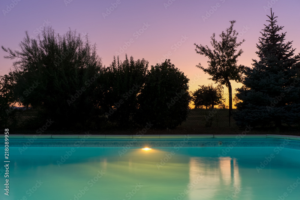 Outdoor blue liner swimming pool with lighting at dusk twilight sunset on a clear evening of summer, France.