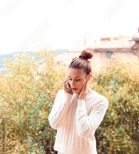 Pretty beautiful young woman in white sweater and underwear, talking on the phone, standing on balcony. Tuscany, Italy. Small depth of field.