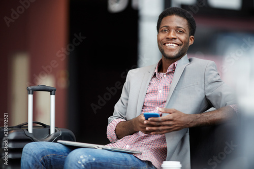 Cheerful adult ethnic man sitting in hall of airport with luggage and holding smartphone while looking at camera