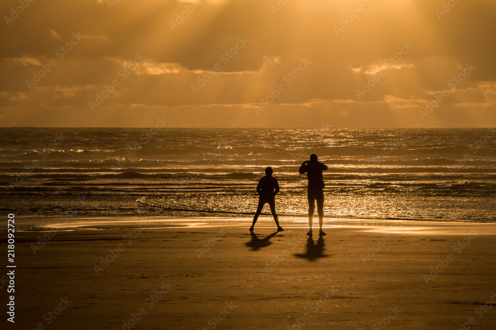 Silhouette image of two people taking photos on the Piha beach, bathed in the Sun, west Auckland, New Zealand