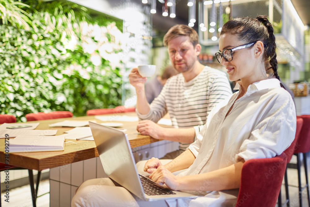 Fototapeta premium Stylish woman using laptop while sitting at table with man having coffee and working together in light office
