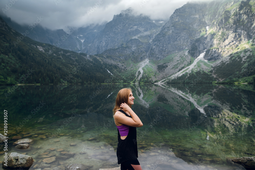 Beautiful young fit woman in sportswear standing on the rocky shore and admiring scenic view of green hills and mountains on Morskie Oko lake, High Tatras, Zakopane, Poland.