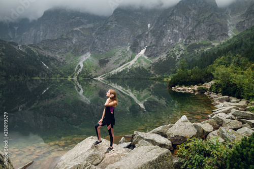 Young beautiful blonde woman in sportswear standing on the rocky shore and enjoying beautiful view of green mountains on Morskie Oko lake, High Tatras, Zakopane, Poland.
