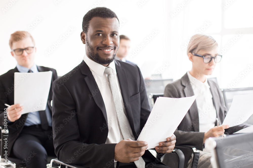 Cheerful African-American man in suit sitting with coworkers in conference hall smiling at camera having business meeting Cheerful African-American man in suit sitting with coworkers in conference hall smiling at camera having business meeting