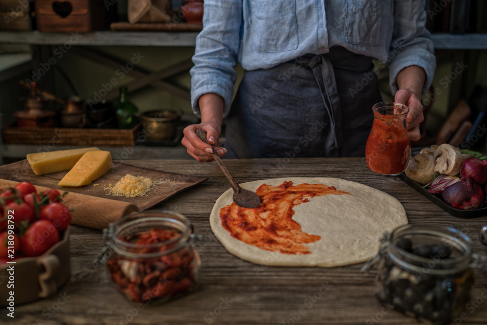 Fresh original Italian raw pizza and ingredients on wooden table, preparation in traditional style. Woman cooking homemade pizza on a wooden table in a home kitchen. Cooking, food and people concept.