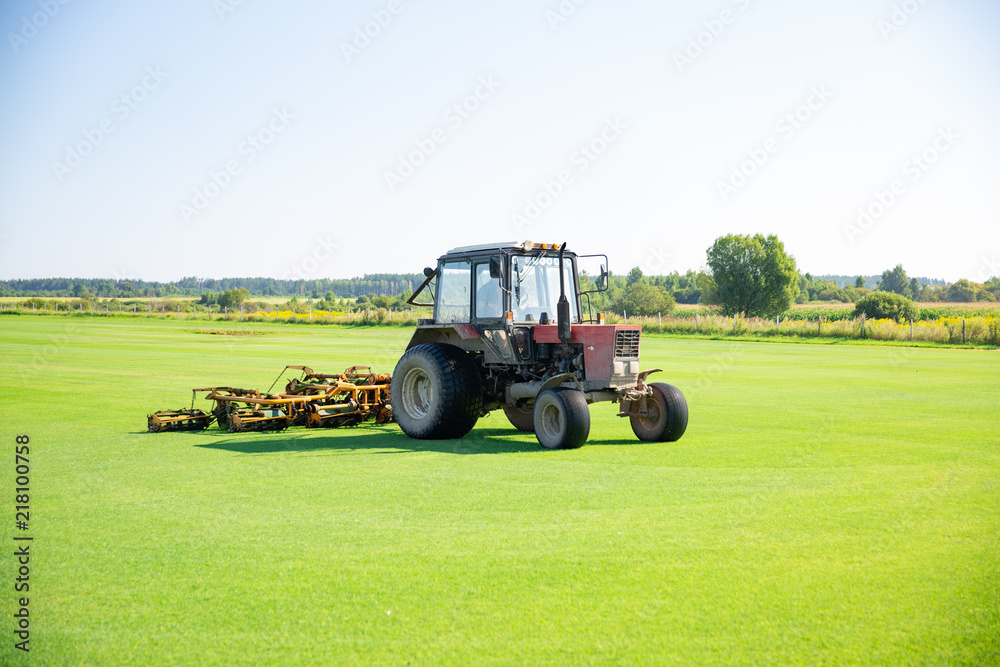 Obraz premium Tractor with a nozzle for care of a field lawn for a horse polo in work. The cut-off grass takes off from mechanical parts of a nozzle. Green flat field. lawn cultivation