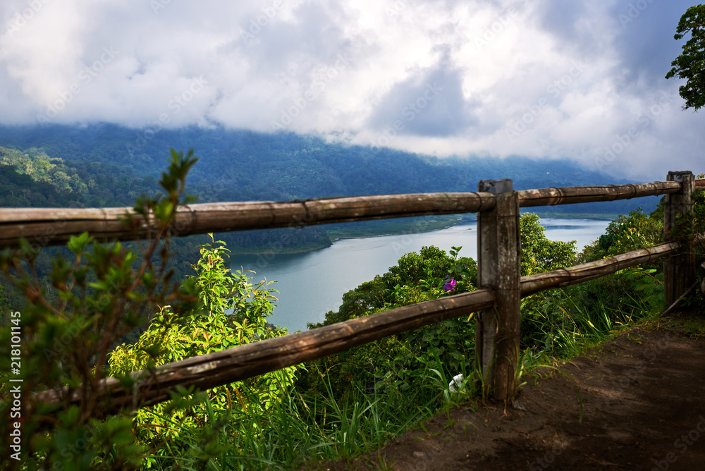 Wooden fence along  the shore of the lake. Low cloud shrouds the distant mountains. Tropical landscape with dramatic clouds in the sky. Lake and mountain view from a hill. Nature background.