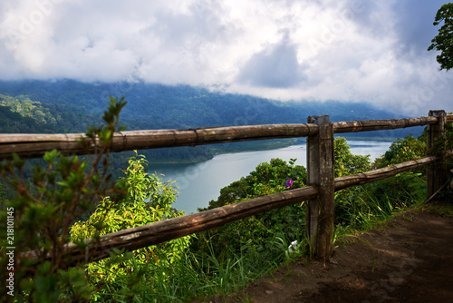 Wooden fence along the shore of the lake. Low cloud shrouds the distant mountains. Tropical landscape with dramatic clouds in the sky. Lake and mountain view from a hill. Nature background.