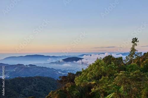 Mountain hill covered with forest on foreground and curly clouds on background with foggy valley with village in between. Mountains during sunrise. Beautiful natural landscape in the summer time.. © eskstock