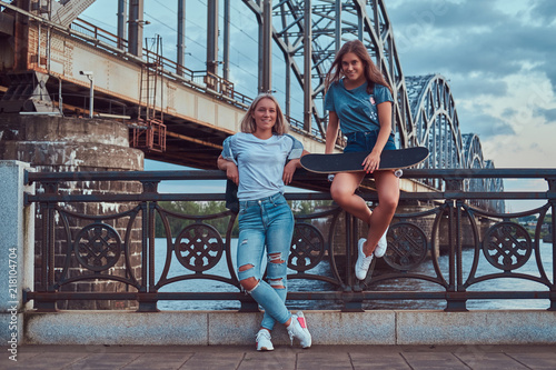 Two happy young hipster girls holds skateboard and leaning on a guardrail on a background of the old bridge.