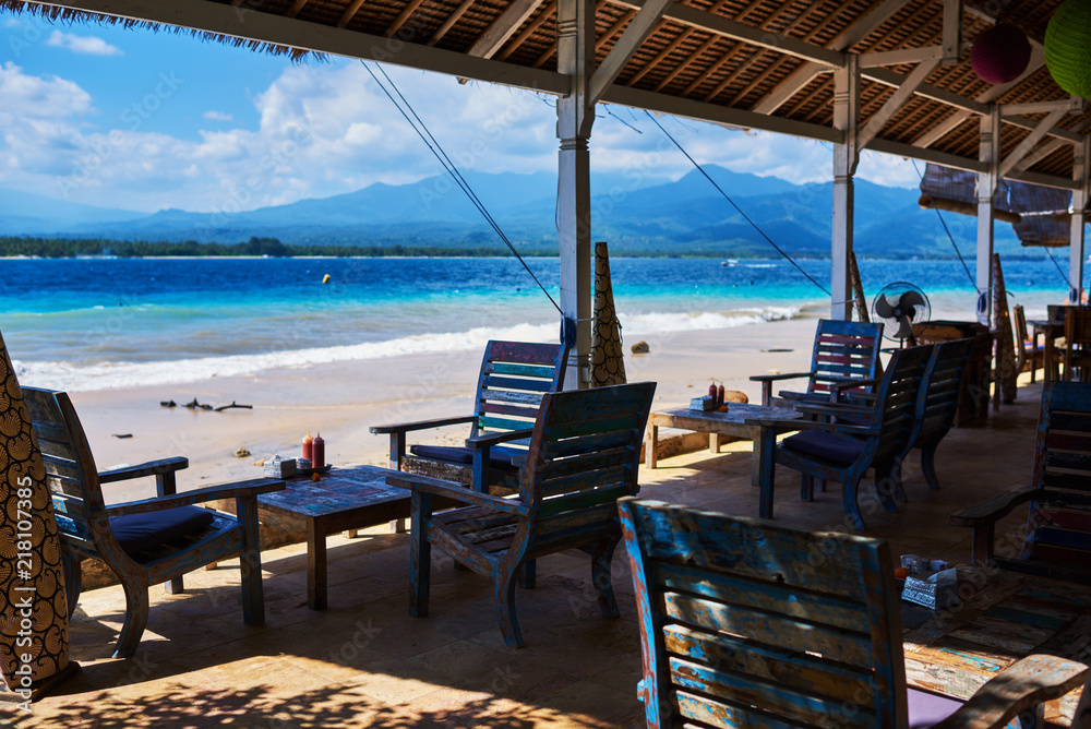 Leisure, travel and tourism concept - close up of table setting at empty open-air restaurant on the paradise beach. Wooden armchairs  on the background of a shining turquoise ocean. Tropical summer.