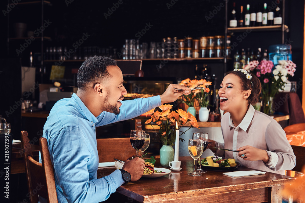 Beautiful African-American couple in love having a great time together at their dating, an attractive couple enjoying each other, handsome man feeding his woman in restaurant.